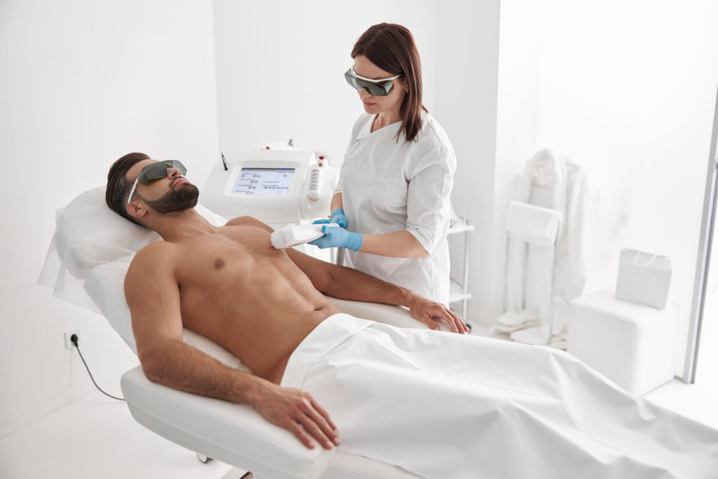 Cosmetologist does laser epilation of chest to athletic man using modern equipment in clinic